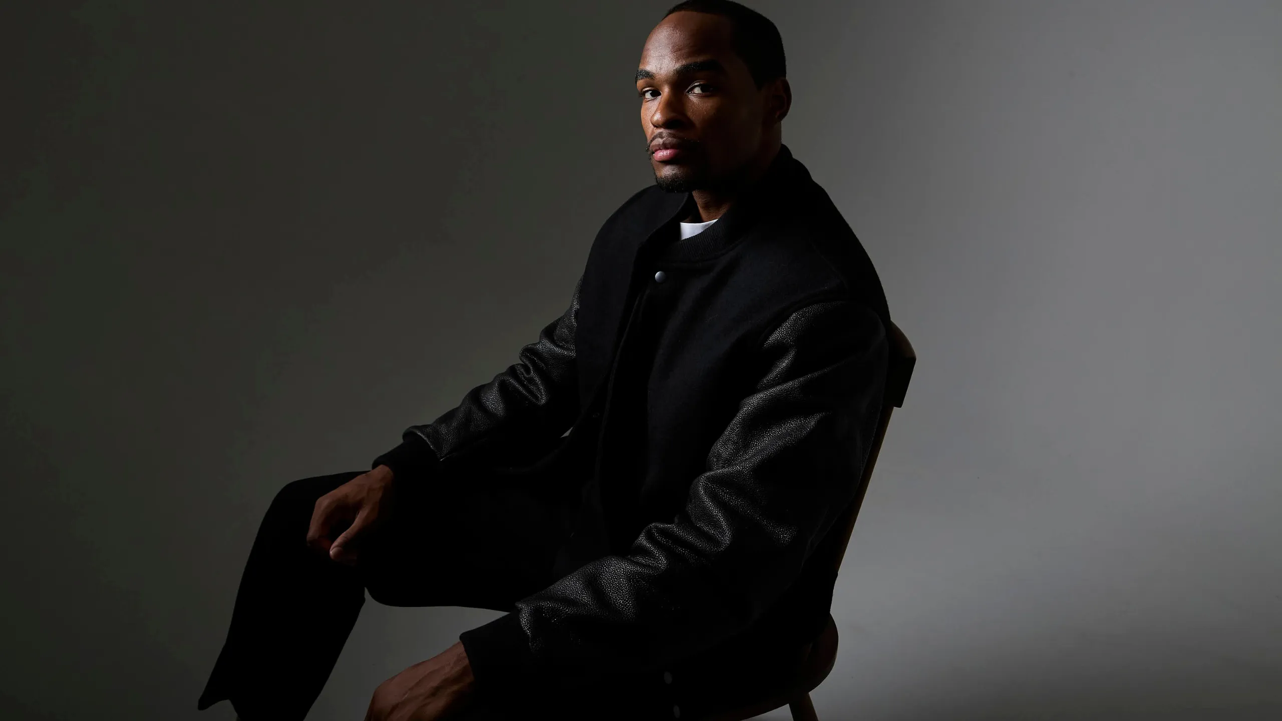 A man seated, wearing a black varsity jacket in Bishu melton wool with sleeves in urushi-lacquered kurozan leather. Ribbing in black. Worn closed, in low light.